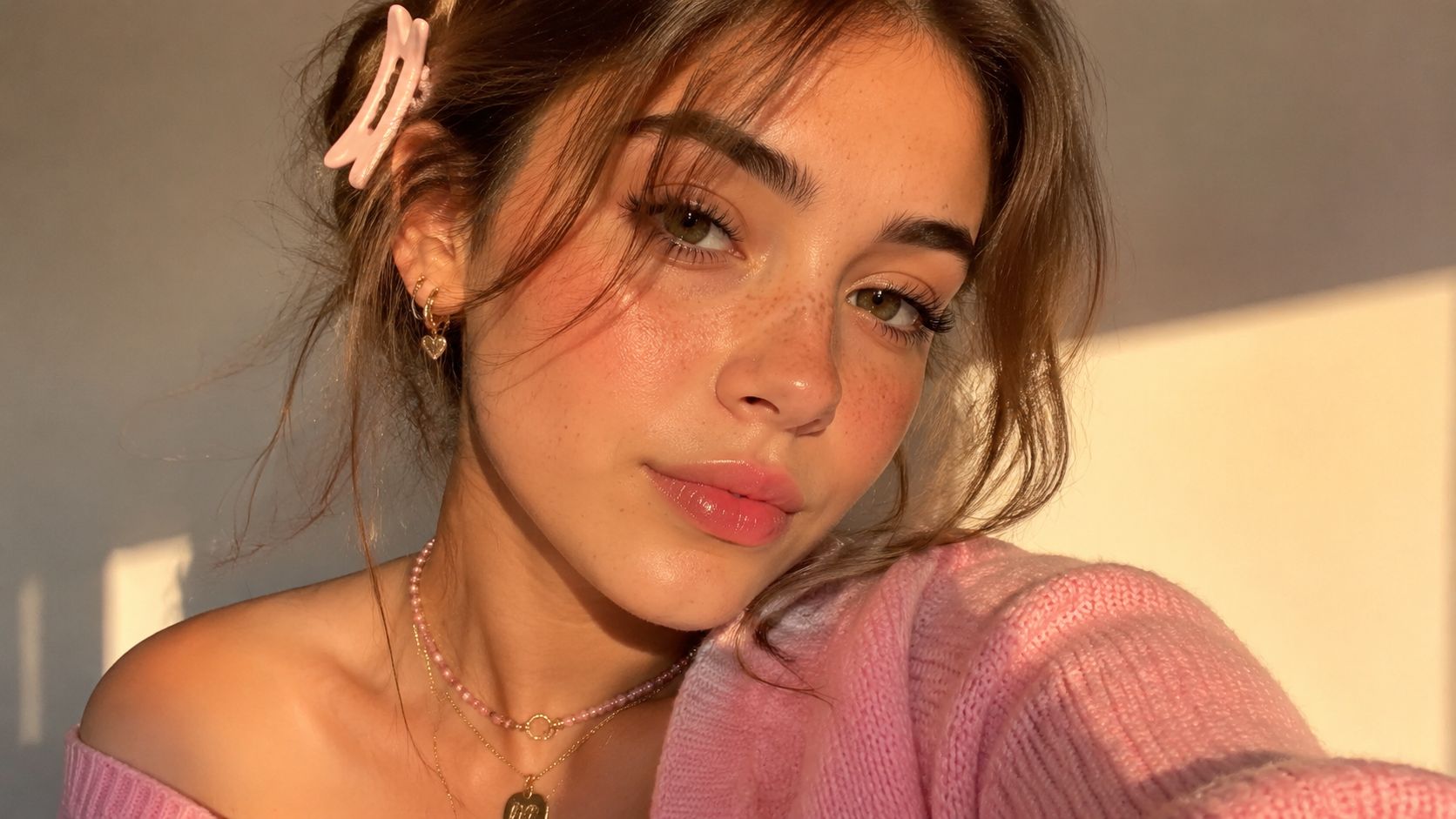 A young woman with a pink sweater and hair clip posing for a selfie in sunlight.