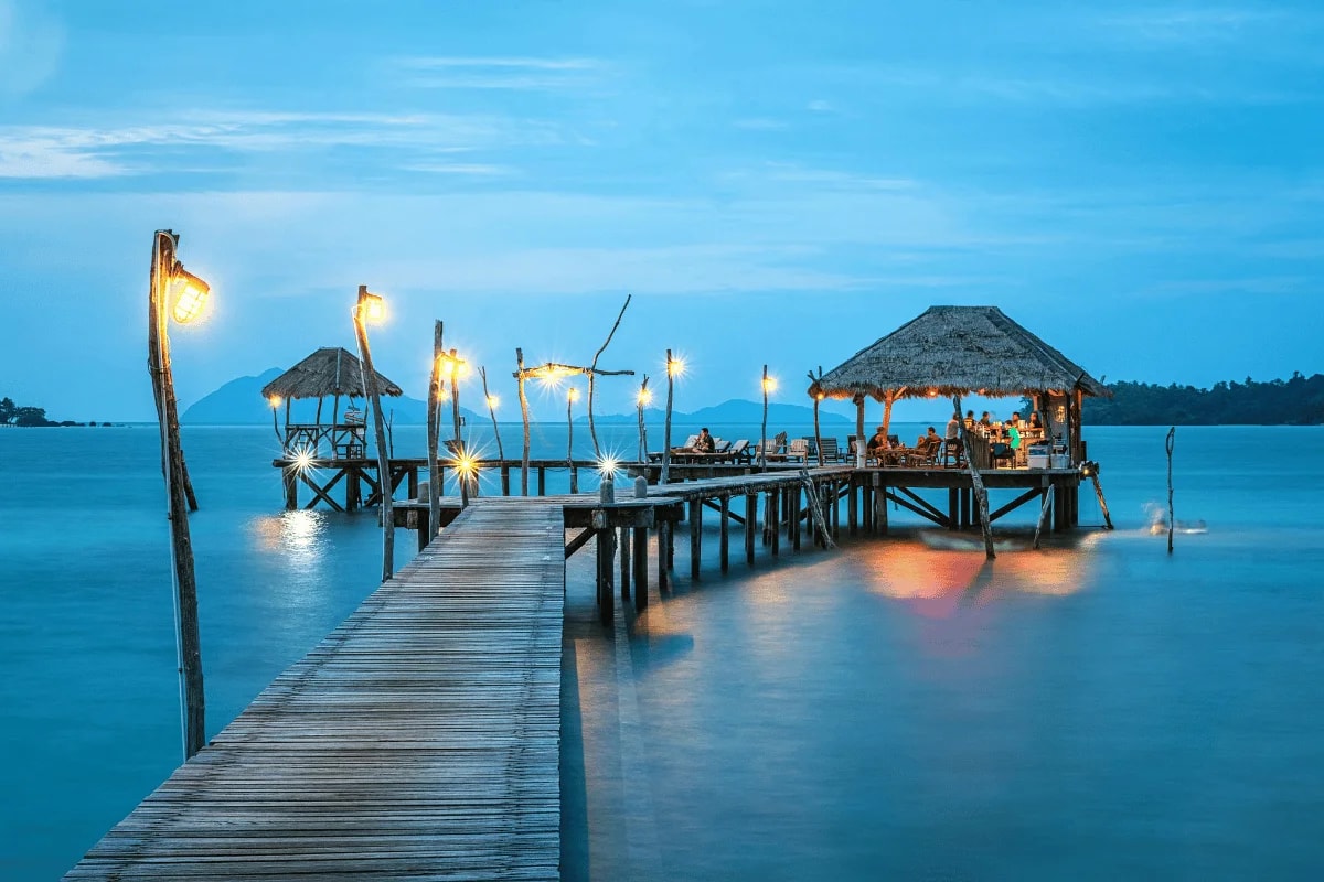 A wooden pier with lit torches leading to a thatch-roofed hut over tranquil waters at dusk.