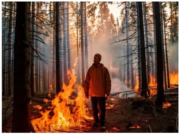 A person stands amidst a forest fire, observing the flames and smoke around.