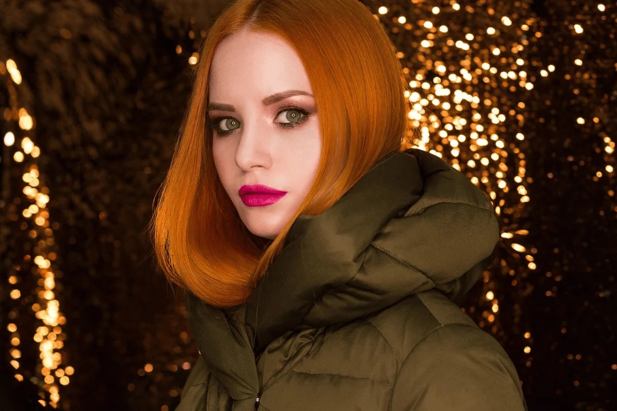 A person with vibrant orange hair wears a stylish olive green puffer jacket against a backdrop of shimmering gold lights.